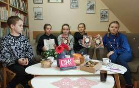 Kilka dzieci siedzi przy stole, trzymają w ręku omawianą książkę, na stoliku logo DKK.