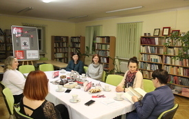 Sześć kobiet siedzi przy stole, jedna z nich czyta książkę, a pozostałe słuchają. W tle regały z książkami. 