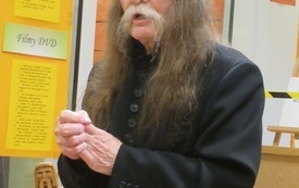 Ubrany na czarno starszy mężczyzna z długimi włosami i wąsami stoi ze splecionymi dłońmi