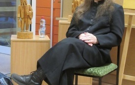 Ubrany na czarno starszy mężczyzna z długimi włosami i wąsami siedzi na krześle