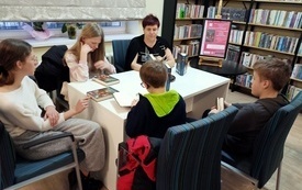Czworo dzieci siedzi przy stole wraz z kobietą, uczestnicy spotkania pochyleni czytają książki. W tle regał z książkami.  