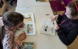 Dwie dziewczynki przeglądają książki z obrazkami przy stole. Na stole znajduje się kilka książek. 