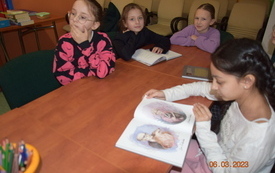 Cztery dziewczynki siedzą przy stole, jedna z nich pochyla się nad książką i ją przegląda. 