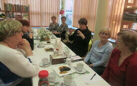 Grupa kobiet siedzi przy stole, przykrytym obrusem, ma stole są też filiżanki, kawa, kwiaty. Jedna z kobiet żywo gestykuluje, pozostałe spoglądają w jej kierunku. 