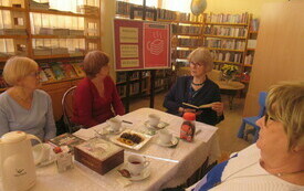 Cztery kobiety siedzą przy stole, kobieta pośrodku trzyma książkę i opowiada pozostałym historię. W tle regały z książkami oraz sztaluga z plakatem Dyskusyjnych Klub&oacute;w Książki. 