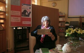Starsza kobieta w okularach z gazetą w ręku. Obok stolik z książkami, zestawem kawowym i bukietem kwiat&oacute;w. 
