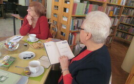 Dwie kobiety siedzące przy stoliku z kawą. Jedna przegląda książką druga patrzy przed siebie.