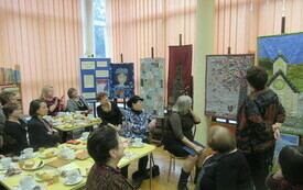 Uczestnicy spotkania oglądające wielkoformatowe obrazy wykonane z tkanin przedstawiające kapliczki, motywy roślinne oraz postać kobiety.