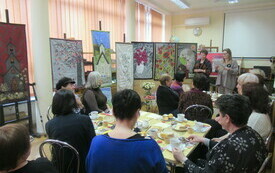 Uczestniczki spotkania siedzące przy stolikach i patrzące w na prace artystyczne wykonane przez prowadzącą spotkanie.