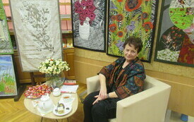 Kobieta w średnim wieku siedząca w fotelu przy stoliku z kawowym na którym stoi bukiet kwiatów, słodycze i herbata. W tle prace W tle obrazy wielkoformatowe przedstawiające kwiaty wykonane z różnokolorowych materiałów.