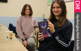 Dwie nastolatki siedzą przy stole, jedna z nich trzyma w rękach książkę, prezentując jej okładkę. 