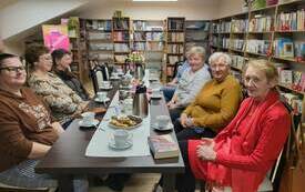 Siedem kobiet siedzi przy długim stole, kilkoro z nich uśmiecha się. W tle regały z książkami. 