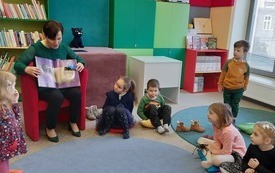 Kobieta siedzi w fotelu, trzyma na kolanach otwarta książkę. Na dywanie siedzą dzieci . 