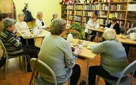 Grupa ludzi siedzi przy kilku stolikach, dwie kobiety rozmawiają. W tle regały z książkami oraz pluszowe maskotki.