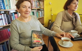 Dwie kobiety siedzą przy stole, jedna z nich prezentuje książkę. W tle za nią regal z książkami. 