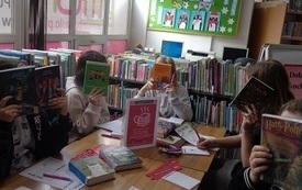 Dzieci siedzą przy stole, zasłaniają twarze okładkami książek. W tle okna i regały z książkami. 