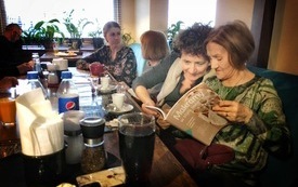 Kobiety na pierwszym planie przeglądają książkę. Pomieszczenie, w którym się znajdują jest przyciemnione.