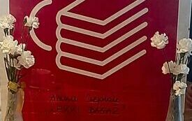 Plakat Dyskusyjnych Klubów Książki, po jego bokach wazony z kwiatami. Prze plakatem dwie książki na stole.