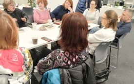 Grupa ludzi siedzi przy stole, kobieta w okularach żywo gestykuluje i m&oacute;wi, pozostałe osoby zwr&oacute;cone są w jej stronę i słuchają 