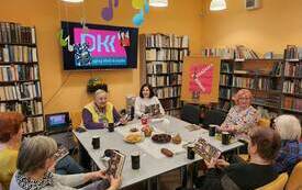 Siedem kobiet siedzi wok&oacute;ł stołu, w tle ekran z logo Dyskusyjnych Klub&oacute;w Książki. 