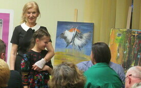 Starsza kobieta obejmuje ramieniem dziewczynkę w kwiecistej sukience. W tle obraz z dwoma ptakami.