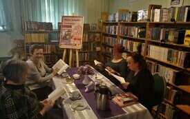 Cztery kobiety siedzą przy stole i trzymają w rękach otwarte książki. W tle regały z książkami. 