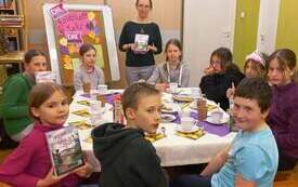 Kilka dzieci siedzi wok&oacute;ł stołu, trzymają książki z ręku i pozują do zdjęcia.