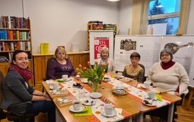 Pięć kobiet siedzi przy stole, na którym znajdują się książki, serwetki, ciastka. W tle na sztaludze plakat DKK.