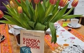 Na stole książka, bukiet tulipanów w wazonie, ciastka. W tle plakat Dyskusyjnych Klubów Książki