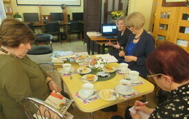 Cztery starsze kobiety siedzą przy stole, na kt&oacute;rym leżą książki, ciasto i filiżanki z kawą