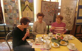 Trzy starsze kobiety siedzą przy stole, na kt&oacute;rym stoją filiżanki z kawą i talerzyki z ciastem