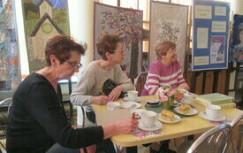 Trzy starsze kobiety siedzą przy stole, na kt&oacute;rym leżą książki, ciasto i filiżanki z kawą