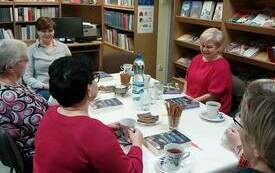 Kilka kobiet siedzi przy stole, rozmawia. Na stole znajdują się filiżanki, ciastka. W tle regały z książkami. lada biblioteczna.