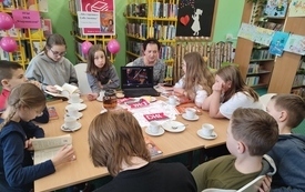 Kilkanaście dzieci siedzi i słucha, przed nimi kobieta z laptopem, na którym coś się wyświetla.