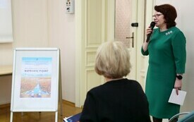 Kobieta w zielonej sukience stoi z mikrofonem w ręku. Obok siedzi druga kobieta i stoi potykacz.