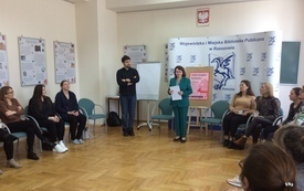 Kobiety siedzą na krzesłach w koło, z przodu kobieta mówi do reszty, obok mężczyzna słucha.