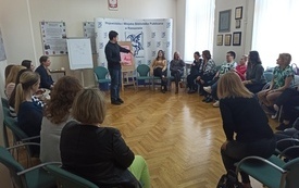 Kobiety w kole siedzą na krzesłach i słuchają mówiącego do nich prowadzącego spotkanie.