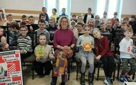 Duża grupa dzieci siedzi na krzesłach, kilka osób trzyma książki. Obok nich siedzi pisarka Joanna Jagiełło.