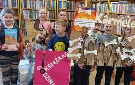 Dzieci trzymają kartonowego psa, napis Karmelek oraz książkę. Obok plakat DKK.