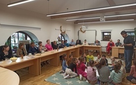 Grupa dzieci siedząca w k&oacute;łeczku na dywanie wpatrzona w stojącego młodego mężczyznę. Wok&oacute;ł przy stolikach siedzą kobiety w r&oacute;żnym wieku. 