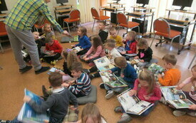 Grupa dzieci siedząca na dywanie. Dzieci trzymają na kolanach książki. Mężczyzna podaje książkę dziewczynce.