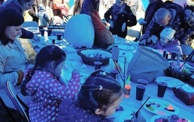 Uczestnicy warsztat&oacute;w skupieni wok&oacute;ł stołu z przyborami do malowania malują kamienie.