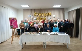 Grupa ludzi stoi, cześć siedzi przed stołem przykrytym obrusem. Obok plansza z plakatem Dyskusyjnego Klubu Książki.