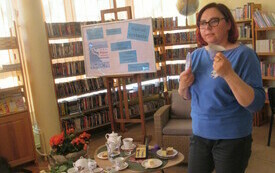 Kobieta w niebieskim sweterku trzyma w ręku filiżankę oraz porcelanowy talerzyk. Obok na stoliku r&oacute;żne porcelanowe naczynia.  W tle regały z książkami.