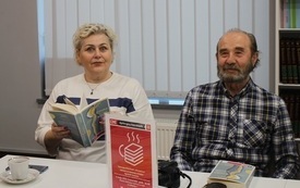 Kobieta i mężczyzna siedzą przy stole, kobieta trzyma książkę. Na stole logo Dyskusyjnych Klubów Książki.