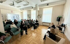 Publiczność zgromadzona na spotkaniu zwr&oacute;cona w stronę dw&oacute;ch kobiet siedzących na fotelach - prowadzącej i gościa spotkania. 