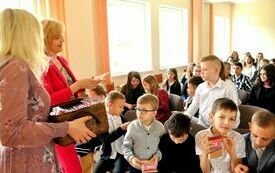 Kilkanaście dzieci siedzi, a przed nimi dwie kobiety jedna z nich mówi a druga trzyma kosz z umpominkami.