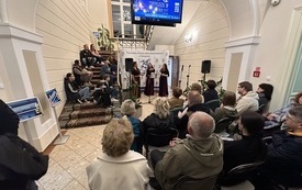 Zgromadzona publiczność siedzi na krzesłach i schodach podczas koncertu trzech kobiet w słowiańskich strojach z zespoły Pełna.