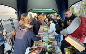 St&oacute;ł z książkami. Wok&oacute;ł stołu zgromadzeni ludzie przeglądają książki.  