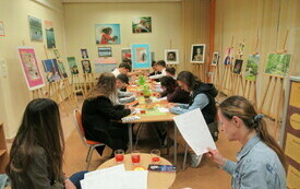 Przy stoliku dwie kobiety, jedna trzyma w ręku kartkę z krzyżówką. Przed kobietami stół przy którym siedzi młodzież i rozwiązuje zadanie konkursowe. Wokół na sztalugach grafiki.